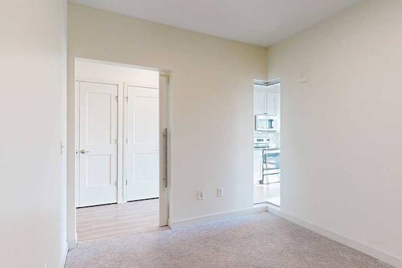 Empty room with beige walls and carpet, featuring a white door leading to another space with wooden floors. A glimpse of a modern kitchen is visible.
