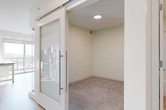 A minimalist room with beige carpet, illuminated by a ceiling light. Frosted glass sliding doors separate it from a bright kitchen with a view.