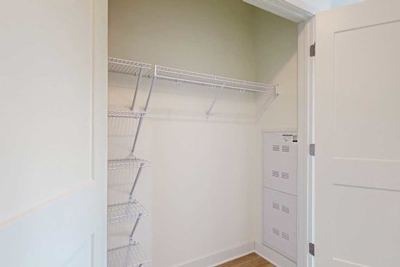 Small, empty closet with white wire shelves on left and top walls, cream-colored walls, wooden floor, and open white door;
