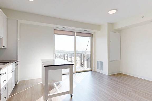 Bright, modern kitchen with light wood flooring, white cabinets, and a central island. Large glass doors lead to a balcony, creating an airy feel.