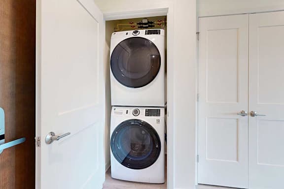 Stacked front-loading washer and dryer in a small laundry closet. The space is neatly organized, with white doors open