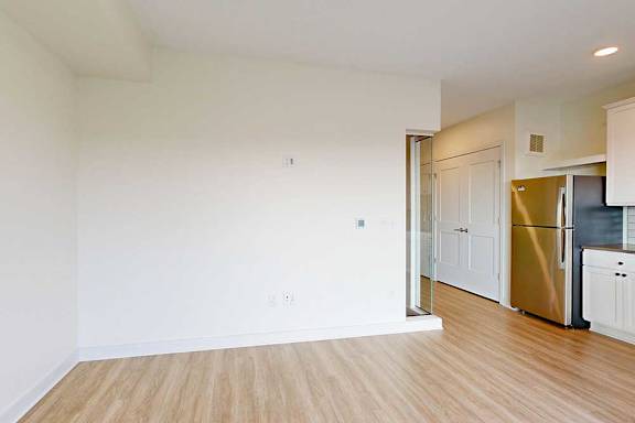 A bright, empty room with light wood flooring, white walls, and a stainless steel refrigerator.
