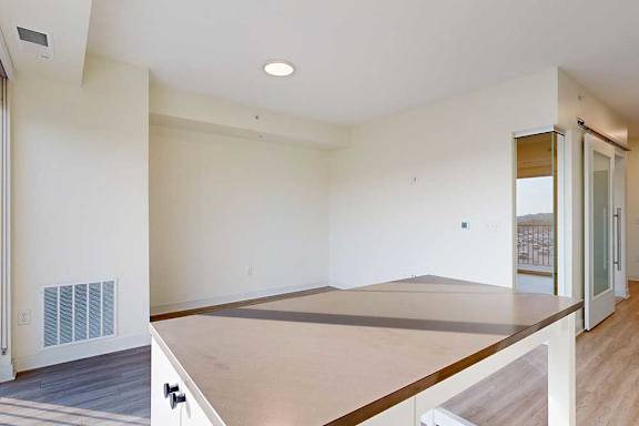 Bright, empty room with light wood flooring and beige walls. A large window lets in sunlight, and a wooden countertop extends into the space.