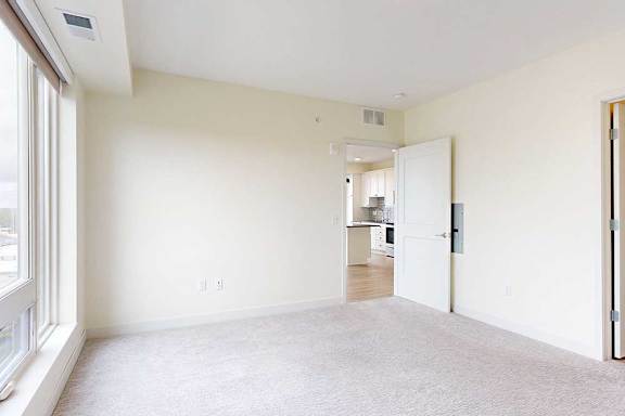 Bright, empty room with beige carpet and off-white walls, large window on the left. An open door leads to a kitchen with wood flooring.