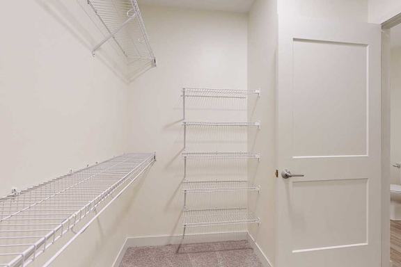 Empty walk-in closet with beige walls and white wire shelving on the left and back walls. A partially open white door is visible
