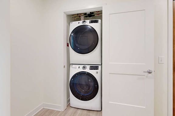 Stacked washer and dryer in a small closet with an open white door, placed on light wooden flooring.