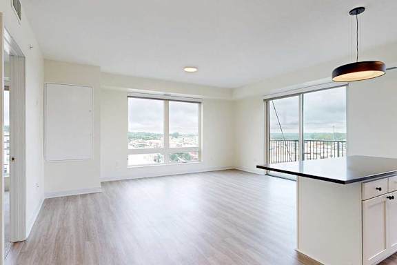 Modern, unfurnished apartment with light wood flooring, large windows showing a city view, white walls, and a black pendant light over a kitchen island.