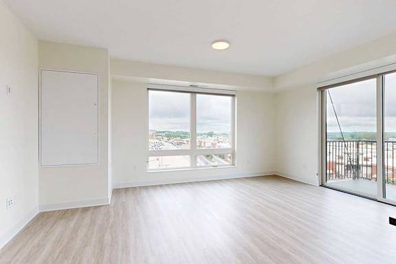 A bright, unfurnished apartment room with light wood floors, large windows showing a cloudy sky and cityscape, and a small balcony, conveying spaciousness.
