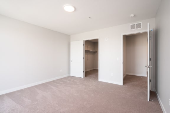 large bedroom with beige carpet and white walls. Two open doors, one leading to the walk in closet and the other leads to the rest of the apartment.