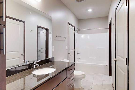 An attached bathroom with a granite sink vanity, dark wooden cabinets, and a tub shower.