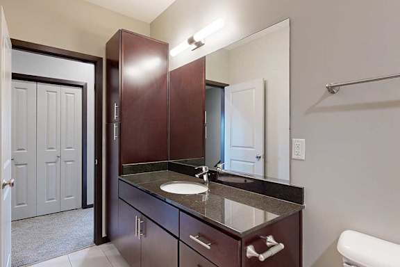 A bathroom with a granite vanity sink, dark wooden cabinets, a tall storage cabinet, and an oversized mirror.
