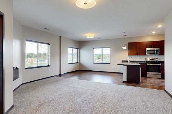A carpeted living room with several windows, open to a dining room and kitchen with hardwood-style flooring.