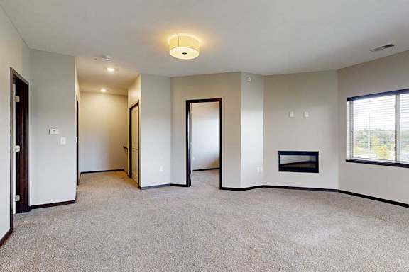 A spacious carpeted living room with an electric fireplace and an open door to a den space.