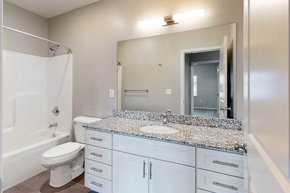 A bathroom with a granite vanity, white wooden cabinets, an oversized mirror with overhead lighting, and a tub shower.