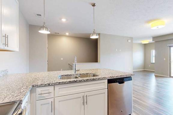 A kitchen peninsula counter with white cabinets, a stainless steel dishwasher, and a double basin sink, in front of a living room.