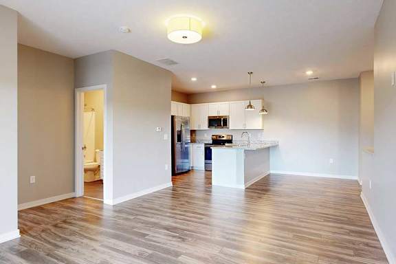 A living room with hardwood-style flooring and kitchen with white wooden cabinets.
