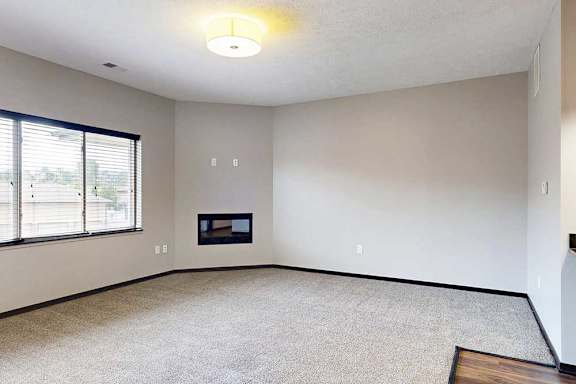 A carpeted living room with an electric fireplace and a large window.