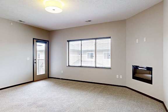 A carpeted living room with a large window, an electric fireplace, and a windowed-door to the balcony.