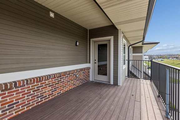 Covered private balcony with wood-look decking, brick accents, and direct access to the apartment entrance.