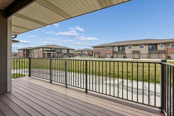 Covered apartment balcony with wood-look decking, black railing, and views of nearby community buildings.