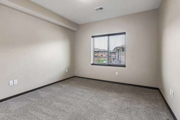 Bright bedroom with gray carpet, neutral walls, and a large window overlooking the apartment community.