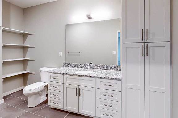 A bathroom with a granite vanity, white wooden cabinets, a tall storage cabinet, an oversized mirror, and built-in linen shelves.