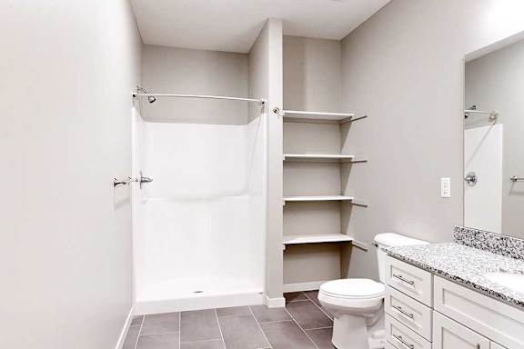 A bathroom with a granite vanity, white wooden cabinets, built-in linen shelves, and a walk-in shower.