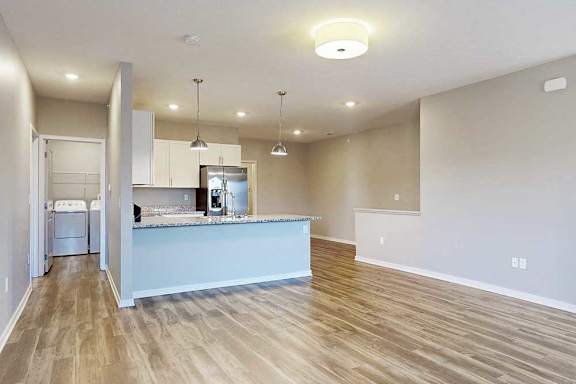 A living room with hardwood-style flooring, open to a kitchen with white wooden cabinets.