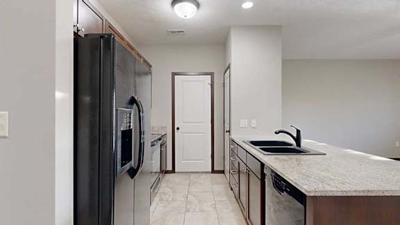 a galley kitchen with granite countertops, dark cabinets, black appliances, a pantry, and a laundry room at the end