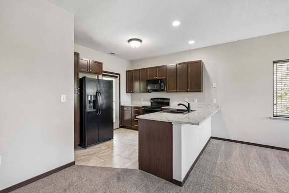 Apartment kitchen with breakfast bar, dark cabinets, black appliances, and carpeted living area nearby.