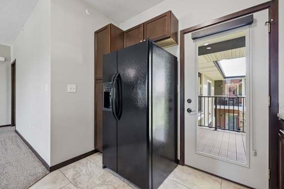 Apartment kitchen entry with black refrigerator, dark cabinets, tile flooring, and glass door to balcony.