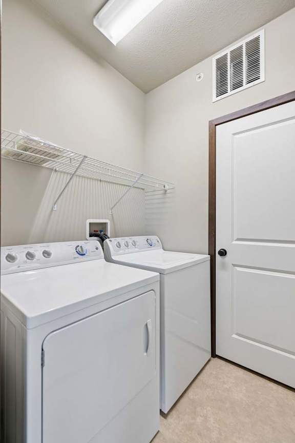 In-home laundry room with white washer and dryer, wire shelving, neutral walls, and tile flooring.