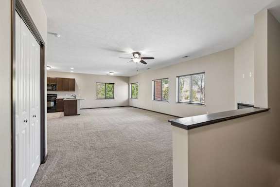 Spacious apartment living room with carpet, ceiling fan, multiple windows, fireplace, and open kitchen.