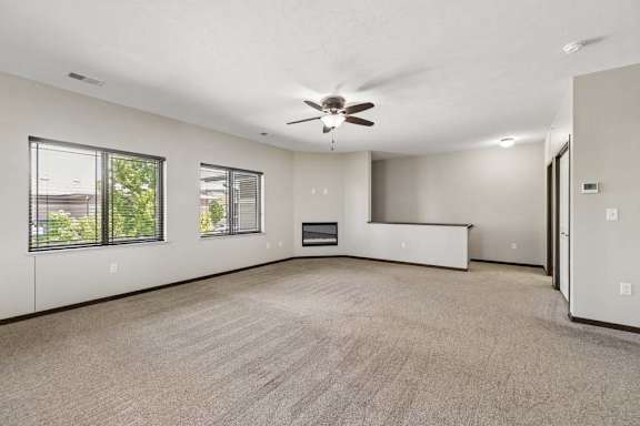 Large carpeted living room with ceiling fan, two windows, corner fireplace, and neutral apartment finishes.