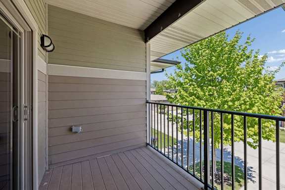 Private apartment balcony with black railing, wood-look decking, and leafy community view.