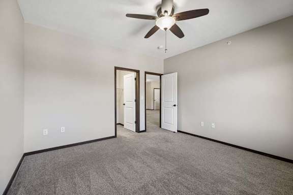 Empty carpeted bedroom with ceiling fan, neutral walls, and doorways to the bathroom and hall.