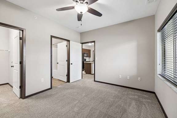 Empty carpeted bedroom with ceiling fan, window with blinds, and doorways to bathroom and kitchen area.