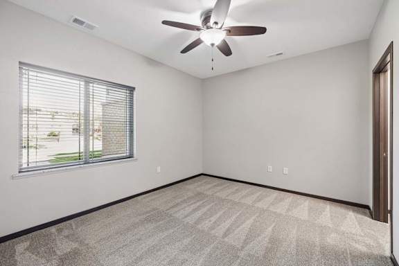 Bright carpeted bedroom with ceiling fan, neutral walls, large window with blinds, and dark trim.