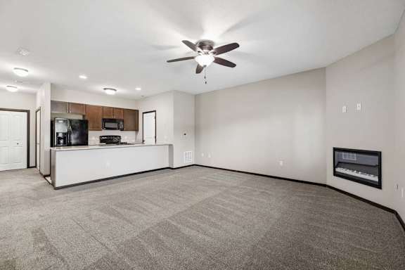 Large carpeted living room with ceiling fan, wall fireplace, window with blinds, and front door.