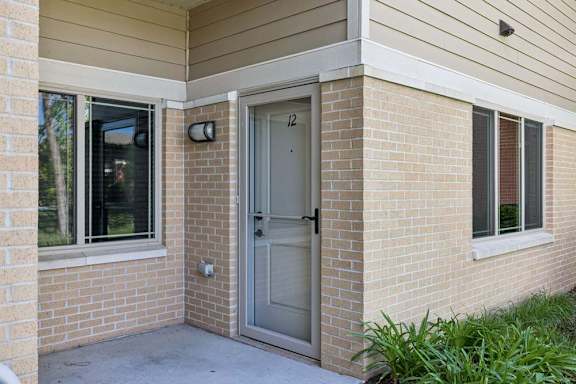 Apartment exterior entry with brick walls, screened front door, large windows, and landscaped greenery.
