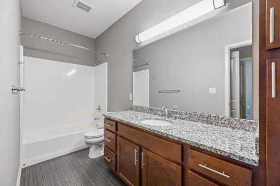 Apartment bathroom with tub and shower combo, speckled countertop, wood vanity, large mirror, and gray tile floor.