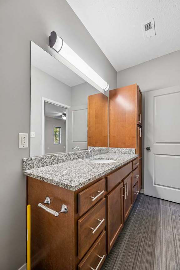 Bathroom vanity with speckled counter, wood cabinetry, large mirror, and bright overhead light.