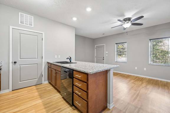 Apartment kitchen island with speckled countertop, wood cabinets, dishwasher, and open living room beyond.