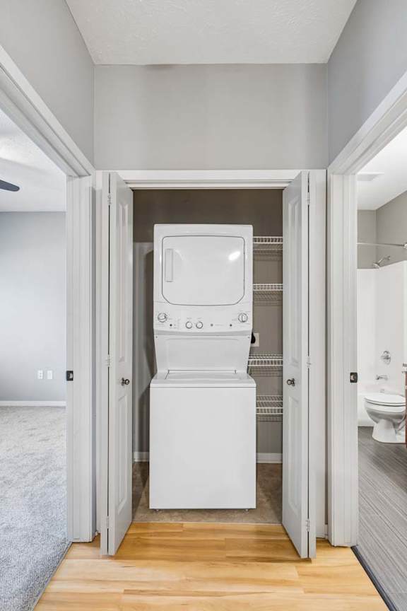 Laundry closet with stacked washer and dryer, wire shelving, double doors, and wood-look flooring outside.