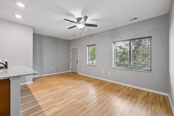 Bright apartment living room with wood-look flooring, ceiling fan, gray walls, and large windows with blinds.