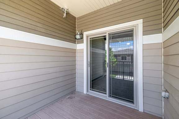 Covered apartment balcony with sliding glass doors, tan siding, plank flooring, and an outdoor wall light.