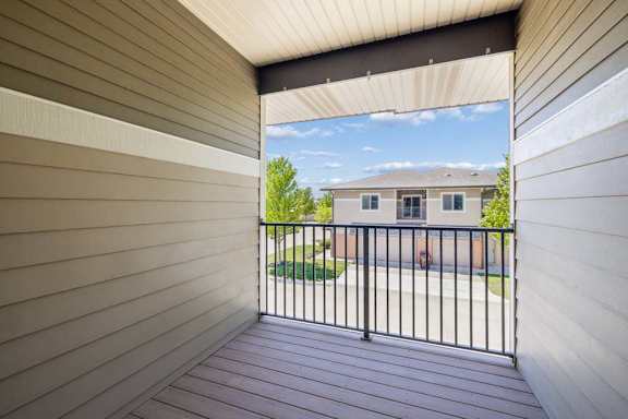 Covered apartment balcony with plank flooring, black railing, tan siding, and a view of neighboring buildings.