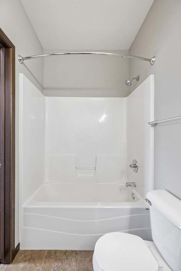 Apartment bathroom with white tub shower combo, curved shower rod, tile flooring, towel bar, and toilet.