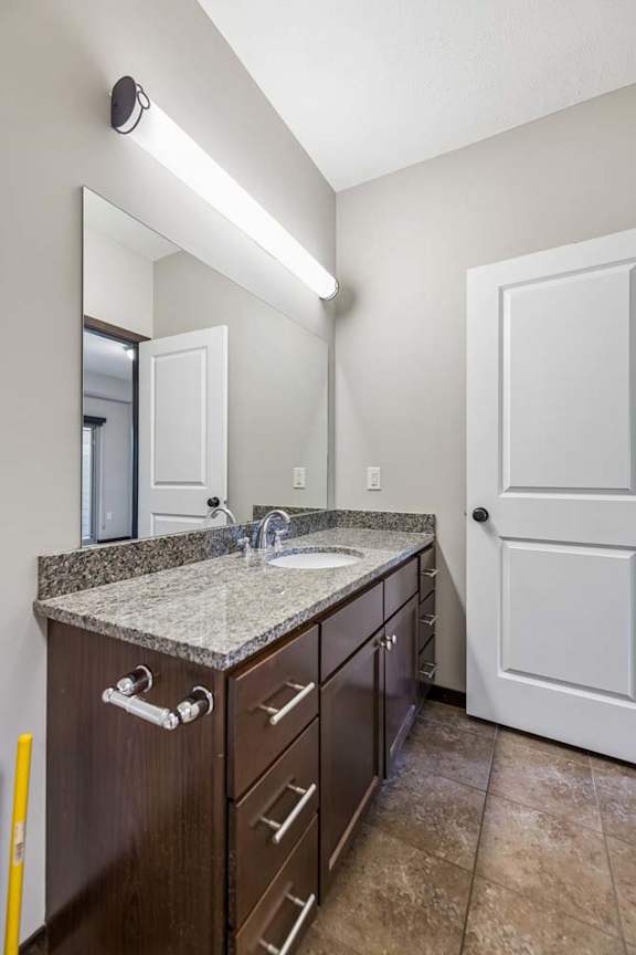 Apartment bathroom with granite-style vanity, dark cabinets, large mirror, tile flooring, and white interior door.
