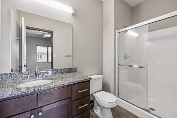 Apartment bathroom with granite-style vanity, dark cabinets, large mirror, toilet, and glass-door shower.
